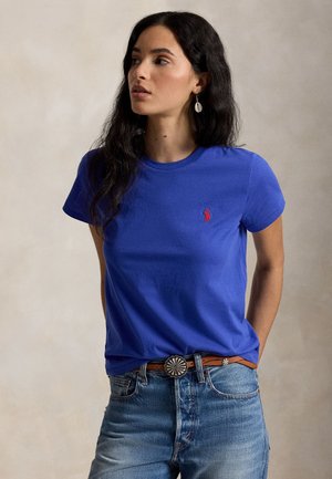 Kobieta z długimi ciemnymi włosami nosi niebieską koszulkę z czerwonym logo, jasnoniebieskie dżinsy i brązowy pasek z srebrną ozdobną sprzączką, patrzy w lewo.