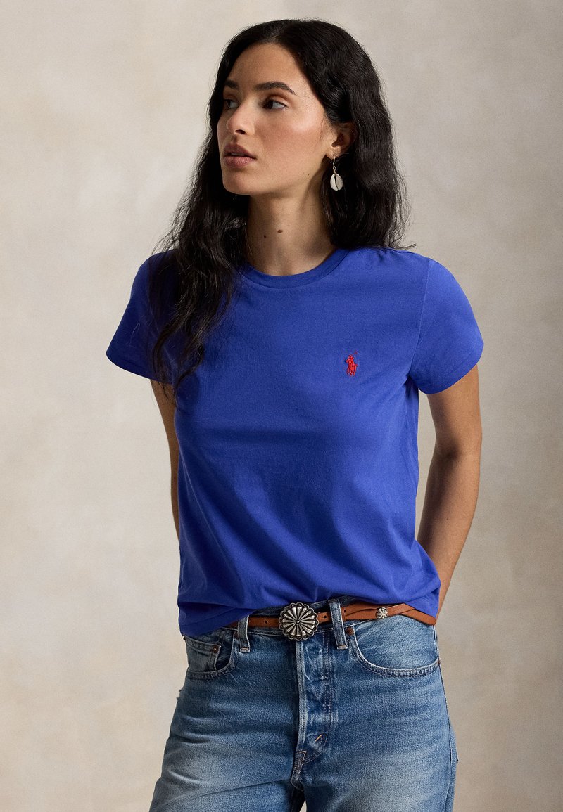 Žena s dlhými tmavými vlasmi má na sebe modré tričko s červeným logom, svetlomodré džínsy a hnedý opasok so striebornou dekoratívnou prackou, pozerá sa doľava.