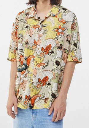 Persona che indossa una camicia a maniche corte con bottoni con motivi floreali arancioni, gialli e beige e pantaloncini di jeans azzurri.