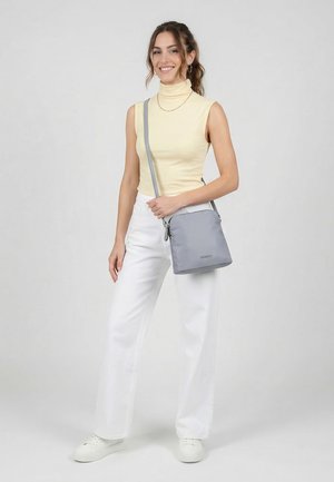 Junge Frau in ärmellosem gelbem Rollkragenpullover und weißer Hose posiert mit grauer Umhängetasche und weißen Turnschuhen vor einfarbigem Hintergrund.