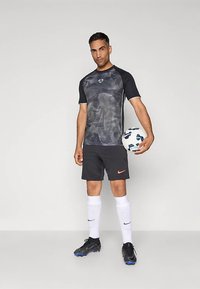 Fekete és szürke rövid ujjú sportpóló hullámmintákkal, fekete rövidnadrággal, fehér zoknival és focicipőkkel; kezében focilabdát tart.