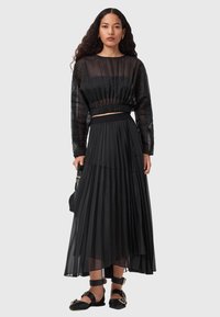 Zwarte gelaagde plissérok met een elastische tailleband en een lange doorschijnende zwarte blouse met geplooide mouwen. Model staat met een tas.