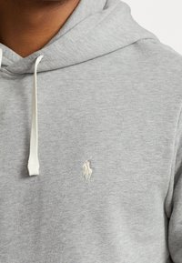 Šedá bavlněná mikina s kapucí, vybavená stahovací šňůrkou, pružnými manžetami a malým bílým logem na hrudi. Měkká textura s uvolněným střihem.