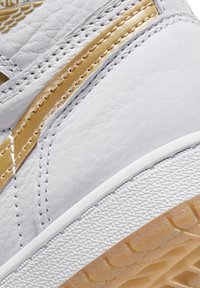 Sneaker in pelle bianca con accenti dorati, superficie testurizzata e suola in gomma. Presenta cuciture morbide e un pattern di battistrada pronunciato.