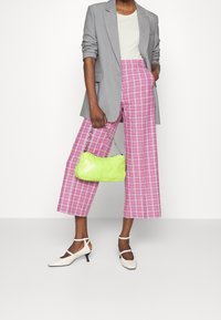 Grijze blazer over een crèmekleurige top, gecombineerd met roze geruite wijde pijpenbroek. Een levendige groene gewatteerde handtas en witte hakken maken de outfit compleet.