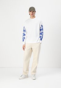 Witte langemouwen shirt met blauwe bloemenontwerpen op de mouwen, gecombineerd met beige broek en witte sneakers. Een grijze pet maakt de look compleet.