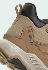 Beige Wanderschuh aus Textil und synthetischen Materialien, strukturierte Oberseite, Gummisohle und Zuglasche; mit schwarzen Fersenakzenten und Branding.