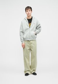 Siva zip-up kapuca z dvema sprednjima žepoma, nošena čez črn majico. Svetlo zelene ohlapne kavbojke in črne čevlje brez vezalk.