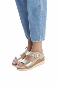 Sandalias de cuña de cuero dorado con dos correas anchas, detalle de hebilla circular y una suela de textura de madera clara. Jeans de mezclilla con dobladillo que complementan.