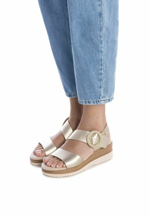 Sandalias de cuña de cuero dorado con dos correas anchas, detalle de hebilla circular y una suela de textura de madera clara. Jeans de mezclilla con dobladillo que complementan.