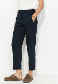 Pantalons navy à coupe droite, avec poches latérales, détails de pli, et légères fentes aux chevilles. Associés à des sabots en suède marron.