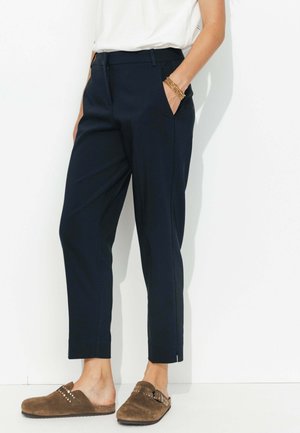 Pantalons navy à coupe droite, avec poches latérales, détails de pli, et légères fentes aux chevilles. Associés à des sabots en suède marron.