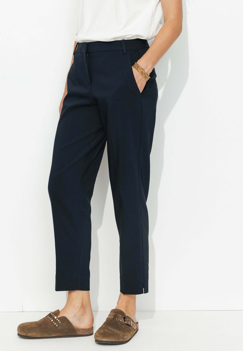 Pantalons navy à coupe droite, avec poches latérales, détails de pli, et légères fentes aux chevilles. Associés à des sabots en suède marron.