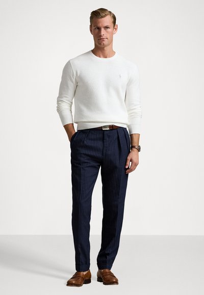 Balts adīti džemperis, tumši zila svītraina bikses, brūns ādas jostiņa un brūnas kurpes. Džemperim ir apaļa apkakle un ribotas aproces.