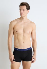 Fekete pamut boxer alsónemű kék derékkal, rajta a "TOMMY HILFIGER" logóval. A modell karjait lazán tartja, ing nélkül áll.