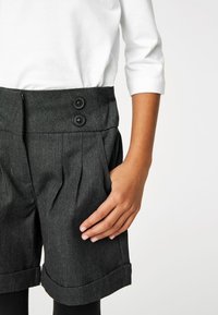 Grijze geplooide shorts met twee zwarte knopen op de tailleband, opgerolde broekspijpen en een gladde textuur. Witte blouse zichtbaar aan de bovenkant.