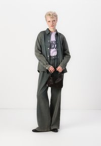 Zelena od jeansa oversized košulja, lavanda grafička majica, široke zelene hlače, drži malu crnu torbicu, crne ravne cipele. Jednostavna bijela pozadina.