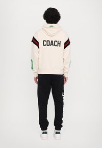 Cream hoodie with black and red stripes, "COACH" text on back; paired with black joggers featuring white logo and green accents.