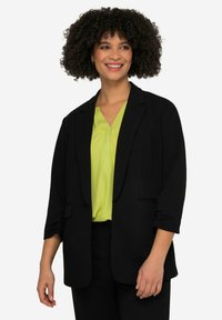 Blazer noir en tissu lisse, avec un col à revers, deux poches avant et des manches trois-quarts, porté sur un chemisier vert vif.