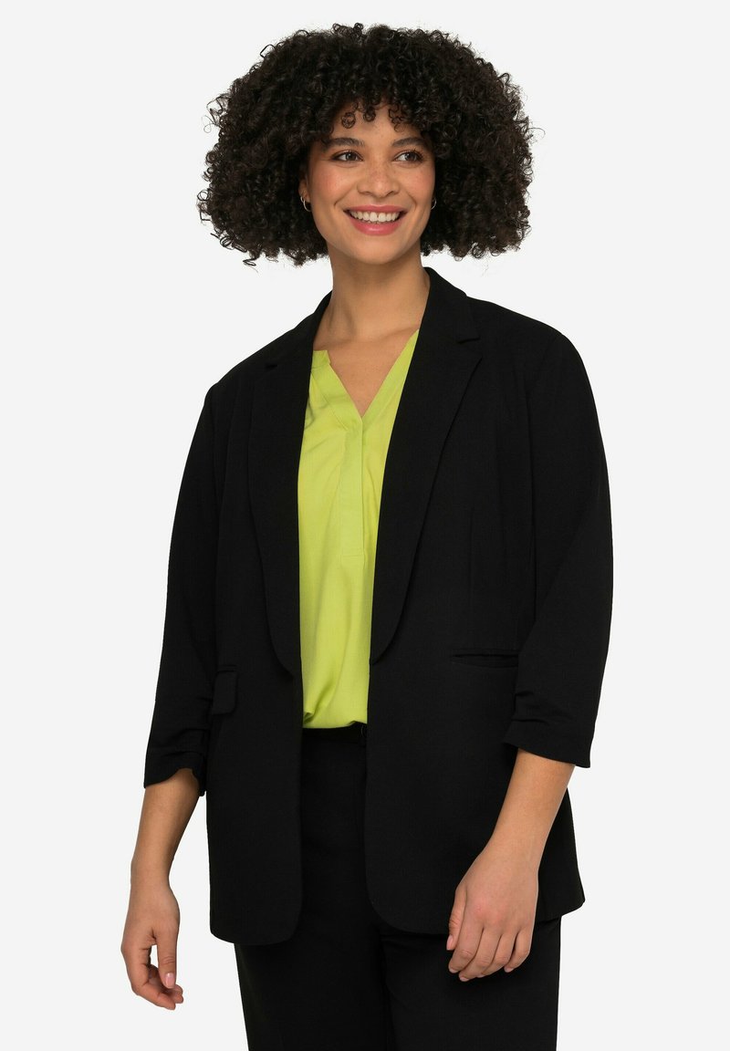 Blazer noir en tissu lisse, avec un col à revers, deux poches avant et des manches trois-quarts, porté sur un chemisier vert vif.