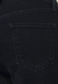 Jean en denim foncé avec une poche arrière en forme de triangle et des coutures contrastées. Le tissu a une texture lisse et un léger éclat.