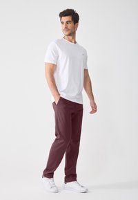 Hvit bomull t-skjorte med korte ermer og et lite gull-emblem, kombinert med bordeauxfargede bukser og hvite joggesko, stående på en nøytral bakgrunn.