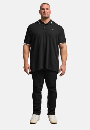 Mand står vendt fremad iført sort polo, sorte bukser og hvide sneakers mod en lys, ensfarvet baggrund.