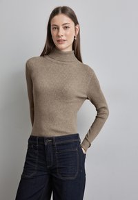 Beiger, gerippter Rollkragenpullover mit langen Ärmeln, tailliertem Schnitt und strukturiertem Finish, kombiniert mit dunkelblauen Jeans mit Eingrifftaschen.