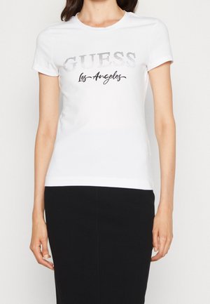 Kobieta ubrana w biały t-shirt z krótkim rękawem ozdobiony srebrnym napisem "GUESS" oraz czarnym napisem "Los Angeles", w połączeniu z czarną spódnicą.