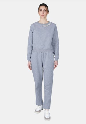 Donna che indossa una felpa grigio chiaro con un piccolo ricamo floreale vicino allo scollo e pantaloni della tuta abbinati, in piedi davanti a uno sfondo bianco.