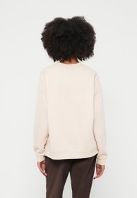 Gaiši bēšs sporta krekls ar apaļu kakla izgriezumu un atkāptiem pleciem, ar brīva piegriezuma siluetu un ribotām aprocēm un apakšu, attēlots no aizmugures.