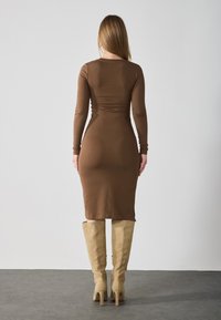 Robe longue marron à manches longues avec une silhouette ajustée et des fentes sur les côtés, associée à des bottes beiges jusqu'aux genoux. Tissu lisse et extensible avec un design minimaliste.