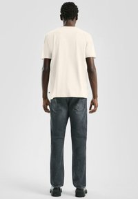 Roomkleurig T-shirt met korte mouwen, gezien vanaf de achterkant, gecombineerd met donkerblauwe spijkerbroek en zwarte schoenen. Simpel ontwerp zonder zichtbare patronen.