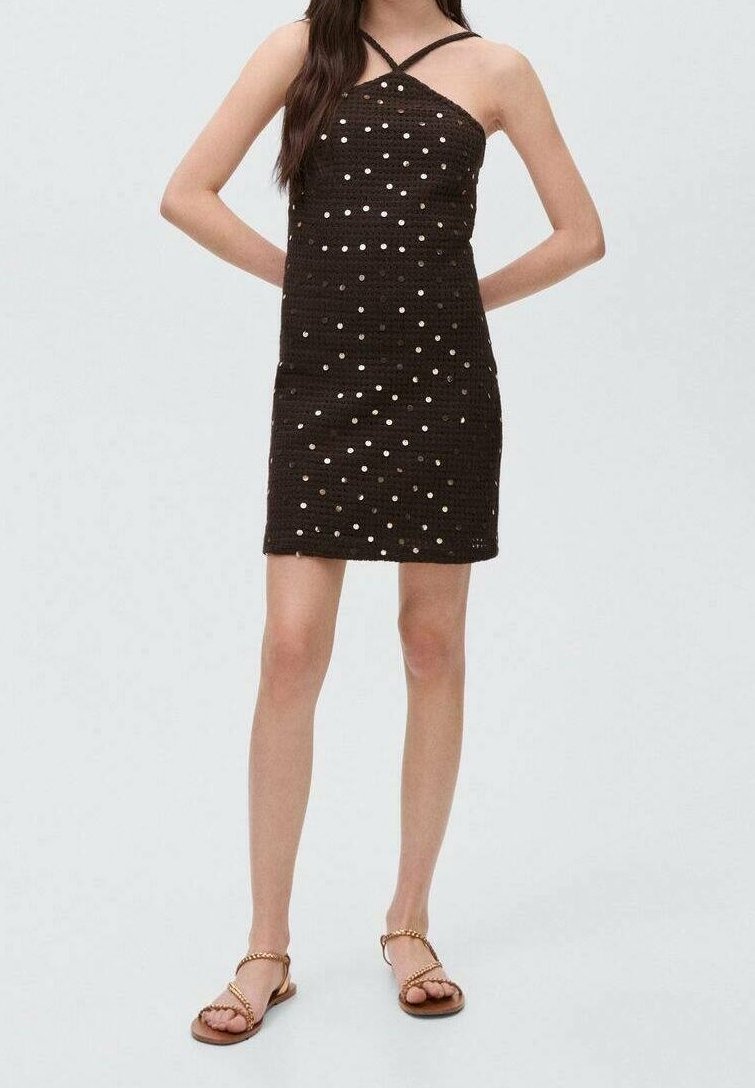 Femme portant une mini-robe noire sans manches à pois blancs avec des bretelles croisées, associée à des sandales plates marron avec des chaînes dorées.