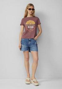T-shirt bordeaux avec un design graphique de soleil, associé à un short en denim bleu clair et des sandales à plateforme. Tenue d'été simple et décontractée.