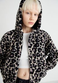 Jakna s kapuljačom u uzorku leoparda napravljena od plišane tkanine. Ima zatvaranje na zip, crne detalje i laganu siluetu s rebrastim rubom.