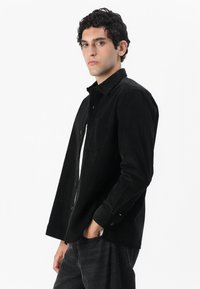 Camisa de pana negra con diseño de botones, un bolsillo en el pecho y mangas largas. Presenta un cuello y una textura sutil.