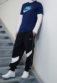 Námořnické tričko Nike s bílým logem, položené přes bílé tričko s dlouhým rukávem. Černé kalhoty s bílými akcenty. Bílé tenisky s průhledn�ými podrážkami.