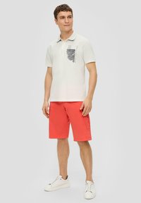 Weißes Poloshirt mit einem Brusttaschendruck, kombiniert mit knalligen Korallenkurzhosen. Das Model trägt weiße Sneaker. Einfaches Design, lässige Passform.