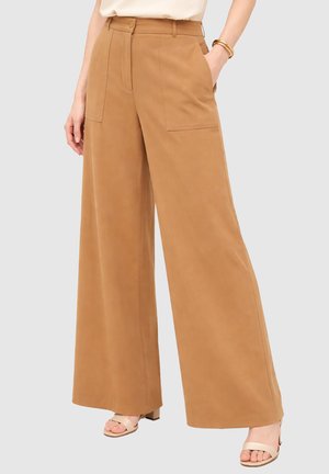 Donna che indossa pantaloni camel a vita alta e gamba larga con tasche applicate frontali, abbinati a sandali beige open toe e braccialetti dorati.