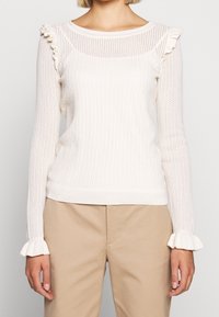 Bílý pletený svetr s dlouhými rukávy a volánky na ramenou a manžetách, doplněný béžovými kalhotami, na stojící osobě.
