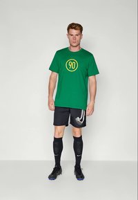 Mand står og har en grøn t-shirt på med gult "90", sorte shorts og sokker med hvide Nike swoosh-designs, samt sorte fodboldstøvler.