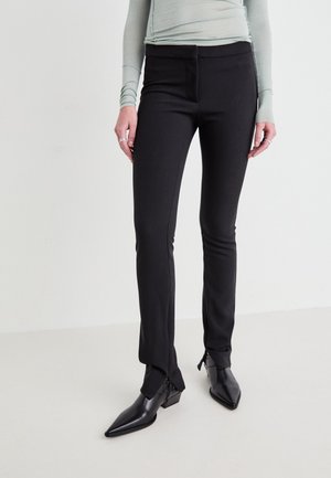 Frau trägt schwarze, schmal geschnittene Hosen mit Reißverschlüssen am Knöchel und schwarze spitze Stiefeletten vor einfarbigem Hintergrund.