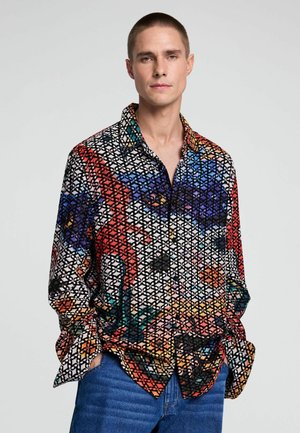 Mann trägt ein farbenfrohes gemustertes Langarmhemd mit geometrischem Motiv, steht mit den Händen in den Taschen vor einem schlichten Hintergrund.