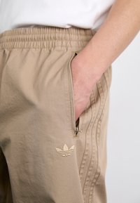 Beige athletic pants with an elastic waistband, zippered side pocket, and embroidered logo. Fabric is smooth and lightweight.
