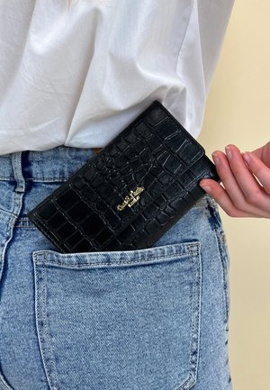 Donna che indossa jeans azzurri chiari e una camicia bianca, mentre infila un portafoglio in pelle nera texturizzata con logo dorato nella tasca posteriore.