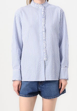 Kvinde iført en langærmet blå og hvid vertikalt striped bluse med flæsekanter og denimshorts mod en ensfarvet baggrund.
