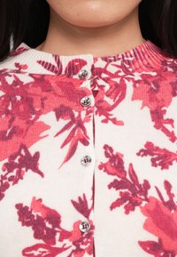 Gros plan d'une personne portant un pull blanc côtelé boutonné avec des motifs floraux rouges et roses autour de l'encolure.