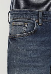Närbild av blå denimjeans med lätt slitage, en myntlåda och gul sömnad, bärs tillsammans med en svart och vit randig skjorta.