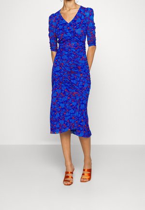 Kobieta w niebieskiej sukience do kolan z czerwonym kwiatowym wzorem, rękawami do łokcia, marszczonymi detalami i pomarańczowymi sandałami na obcasie z odkrytymi palcami.